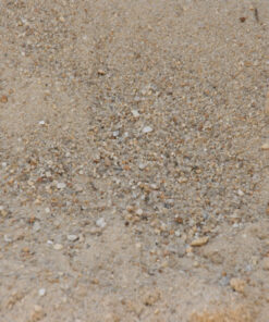 An overhead, close-up photo of a pile of coarse sand. The sand is a light tan and contains small, darker grains and tiny white pebbles. The surface is uneven and has a loose, grainy texture.
