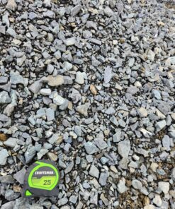 An overhead shot of a pile of gray crushed stone. The rocks are a mix of light and dark gray, with some brown and tan pieces mixed in. A green and black "CRAFTSMAN 25'" tape measure is in the bottom left corner, providing a sense of scale for the size of the rocks.