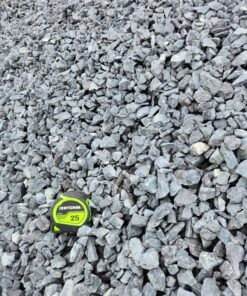 An overhead photo of a pile of gray crushed stone. A green and black "CRAFTSMAN 25'" tape measure is in the bottom center, providing a sense of scale. The rocks are irregular and angular, and they appear to be a mix of light and dark gray tones.