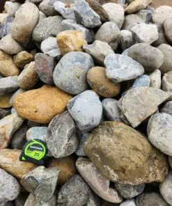 An overhead photo shows a pile of large, rounded river rocks. The rocks are a variety of sizes and colors, including grays, tans, browns, and some with a reddish tint. A green and black "CRAFTSMAN 25'" tape measure is in the bottom left corner, providing a sense of scale for the large rocks.