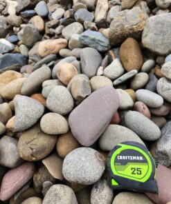 An overhead photo shows a pile of large, rounded river rocks or stones. The rocks are of varying sizes and a mix of natural colors, including gray, brown, and tan. A green and black "CRAFTSMAN 25'" tape measure is in the bottom right corner, providing a sense of scale for the size of the rocks.