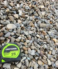 An overhead photo of a pile of rounded river rock gravel, with a mix of colors including gray, brown, and tan. A green and black "CRAFTSMAN 25'" tape measure is in the bottom left corner, providing a sense of scale.