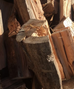 A close-up shot of a pile of freshly chopped firewood. The pieces are of various sizes and shapes, with some showing jagged, splintered edges. The wood is a mix of light tan and dark brown, with visible grain and texture. Sunlight is shining on the pile, casting shadows and highlighting the rough surfaces.