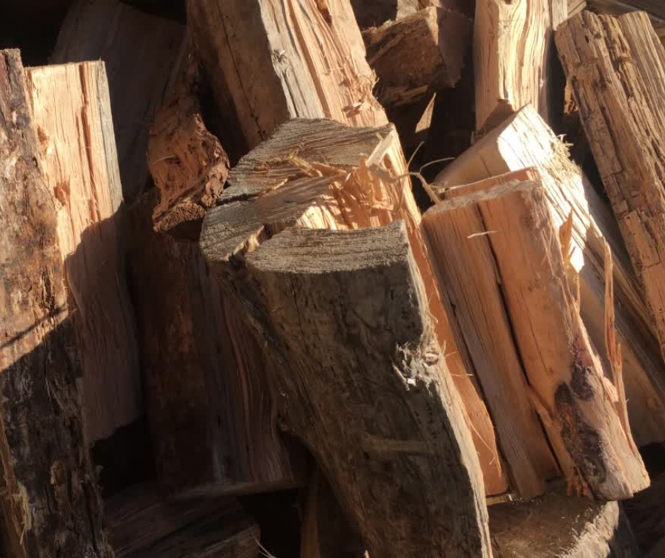 A close-up shot of a pile of freshly chopped firewood. The pieces are of various sizes and shapes, with some showing jagged, splintered edges. The wood is a mix of light tan and dark brown, with visible grain and texture. Sunlight is shining on the pile, casting shadows and highlighting the rough surfaces.