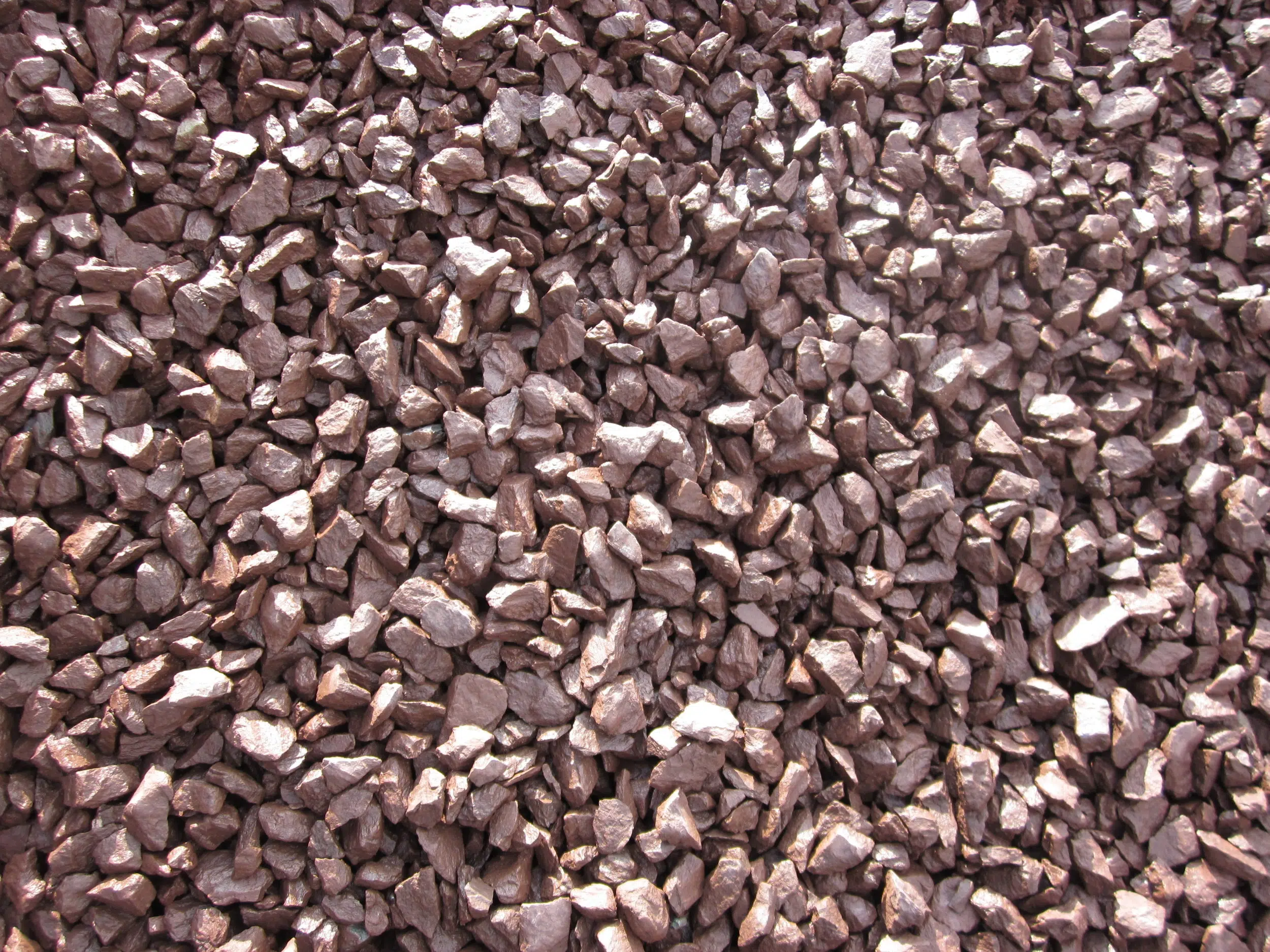 An overhead, close-up photo of a pile of small, reddish-brown crushed stones or gravel. The rocks are irregular and angular in shape, and the sunlight creates a slight sheen on their surfaces.