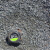 An overhead photo shows a pile of dark gray crushed stone or grit. A black and green "PITTSBURGH" tape measure is in the bottom left corner, providing a sense of scale. The rocks are irregular and angular, with some dirt mixed in.