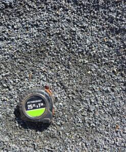 An overhead photo shows a pile of dark gray crushed stone or grit. A black and green "PITTSBURGH" tape measure is in the bottom left corner, providing a sense of scale. The rocks are irregular and angular, with some dirt mixed in.
