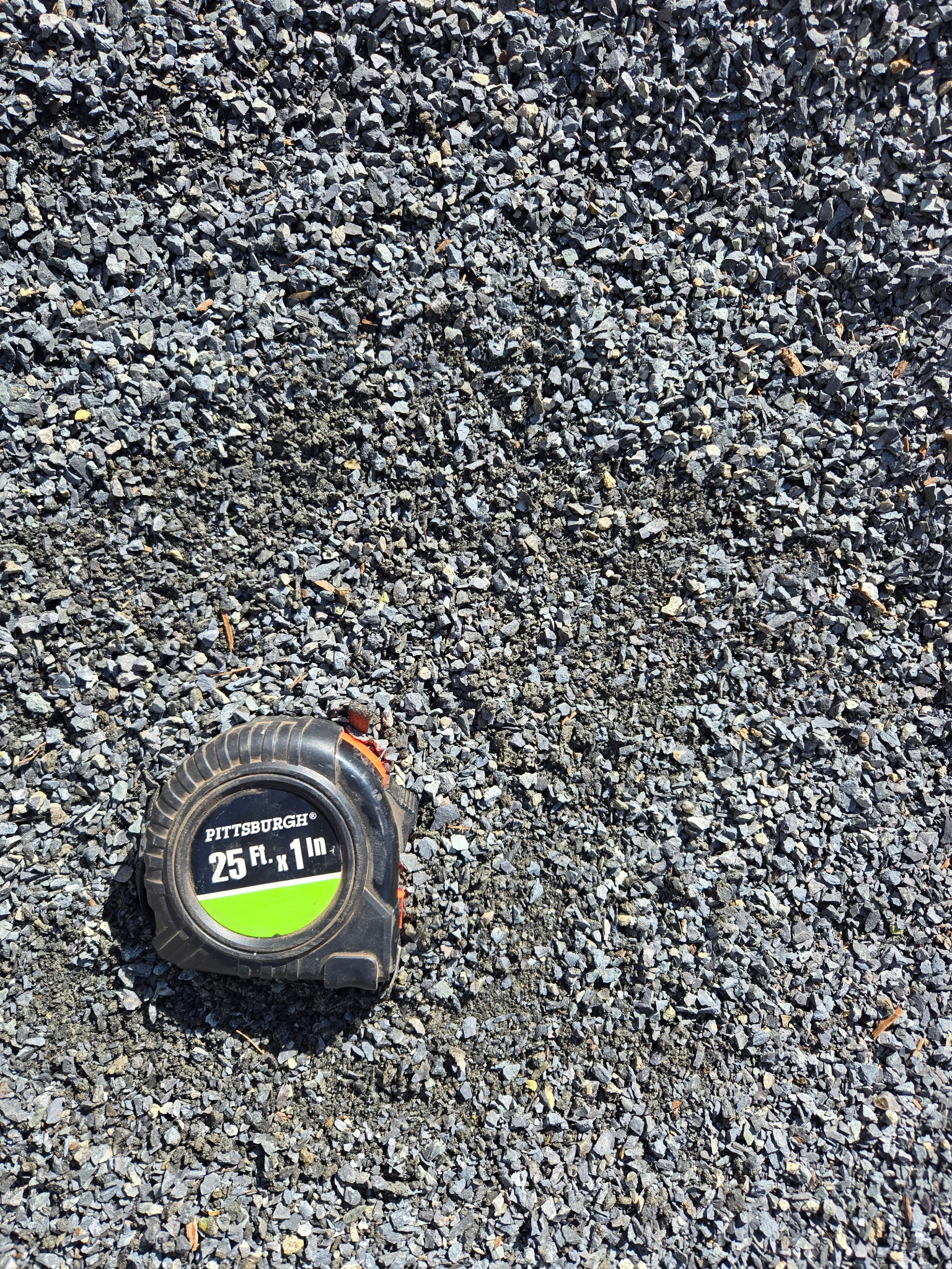 An overhead photo shows a pile of dark gray crushed stone or grit. A black and green "PITTSBURGH" tape measure is in the bottom left corner, providing a sense of scale. The rocks are irregular and angular, with some dirt mixed in.